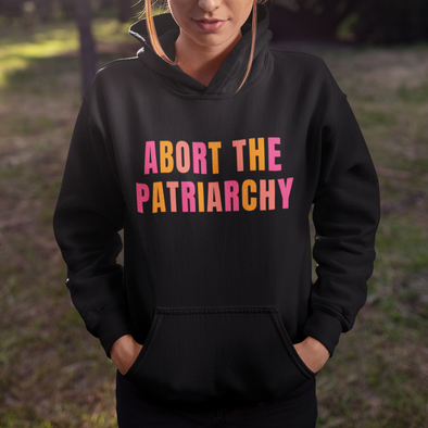 woman standing outside wearing a black hoodie that says abort the patriarchy in pink and orange font
